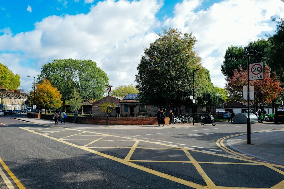 A street scene on Thornton Heath Road showing a small public garden area surrounded by a low red brick wall, with several large deciduous trees exhibiting green and some autumnal reddish leaves. The g